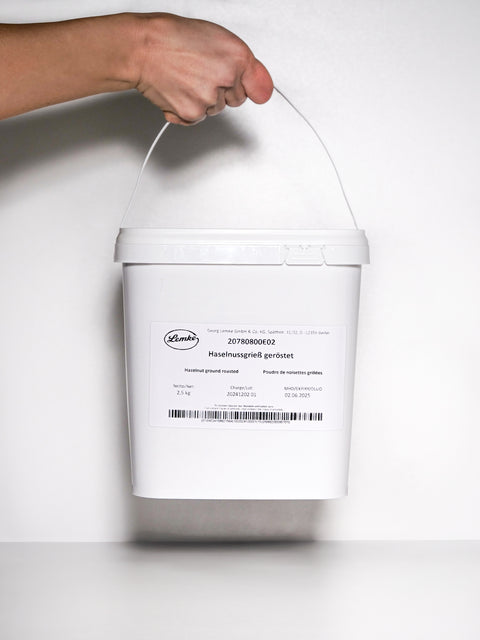 A white bucket labeled 'Haselnussgrieß geröstet,' containing 2.5kg of roasted hazelnut meal, held by a hand gripping the plastic handle. The label includes product details such as net weight, charge lot, and expiration date, with branding from 'Lemke.