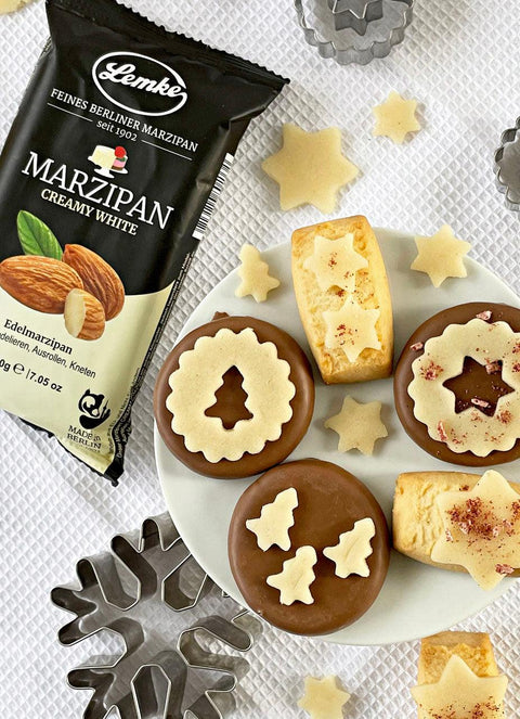 Lemke Marzipan Creamy White 200g Packung neben weihnachtlich dekorierten PlŠtzchen. Die PlŠtzchen sind mit ausgestochenen Marzipan-Sternen und TannenbŠumen verziert, platziert auf einem wei§en Teller mit Backfšrmchen und weiteren MarzipanstŸcken