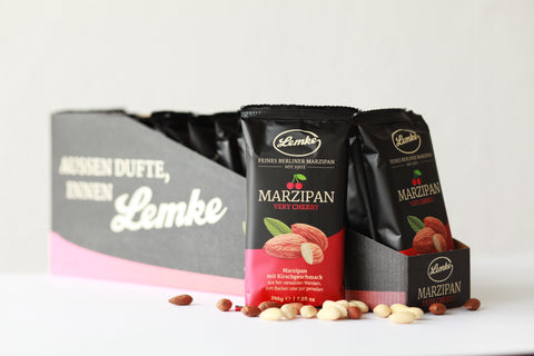 Lemke Marzipan Very Cherry mit Kirschgeschmack, prŠsentiert in einem Tray mit 14 Packungen ˆ 200g. Auf der Vorderseite des schwarzen Trays ist die Aufschrift ãAussen Dufte, Innen LemkeÒ sichtbar. Einige ganze und geschŠlte Mandeln liegen verstreut auf dem Tisch.