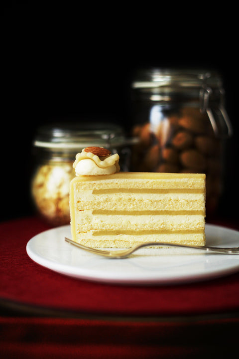 "Ein verfŸhrerisches StŸck Marzipantorte mit mehreren Schichten Biskuit und einer cremigen Sahnehaube,
gekršnt mit einer Mandel. Im Hintergrund stehen GlasbehŠlter mit Marzipankartoffeln und weiteren Mandeln bereit."