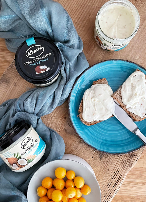 Ein Glas Lemke Stadtstreicher Crunchy Kokoscreme, aufgetragen auf zwei Scheiben Brot, serviert auf einem blauen Teller. Ideal als cremiger und knuspriger Aufstrich für Brot oder Brötchen, begleitet von frischem Obst.
