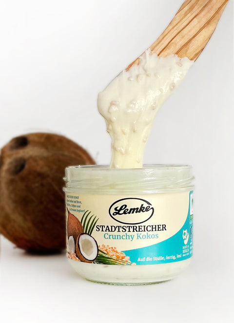 Ein offenes Glas Lemke Stadtstreicher Crunchy Kokoscreme, mit einem Holzlöffel, der die knusprige und cremige Konsistenz zeigt. Perfekt als Topping für Smoothies oder für die Zubereitung von veganen Gerichten.
